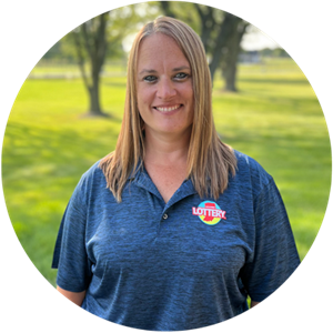 A smiling woman with long hair stands outdoors, wearing a blue polo shirt with a lottery logo, against a green background.